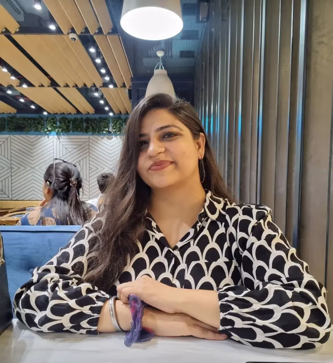 Woman with long dark hair sitting at a table in a modern restaurant wearing a black and white patterned blouse.