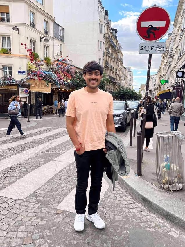 Smiling man wearing a peach t-shirt and black pants standing on a cobblestone street in a city, holding a jacket with buildings and pedestrians in the background.
