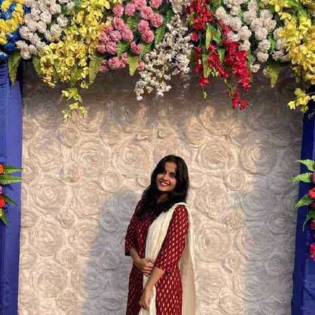 Smiling woman in a red dress with a white scarf posing in front of a floral wall backdrop decorated with colorful hanging flowers.